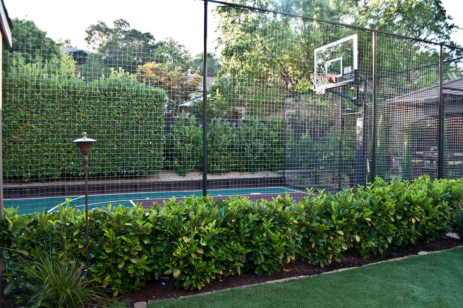 Fenced private basketball court with green border bushes next to manicured lawn