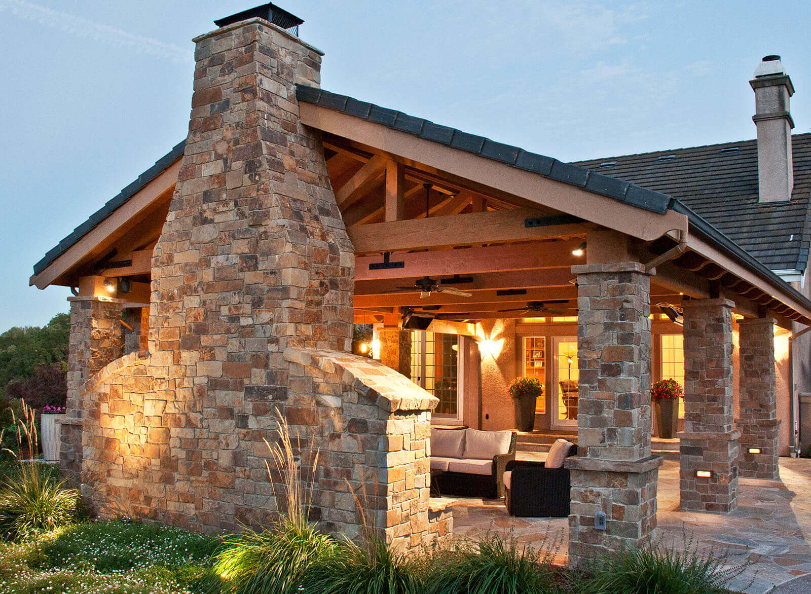 Outdoor warmly lit roofed lounging patio with stone tile columns and fireplace with view of yard behind it