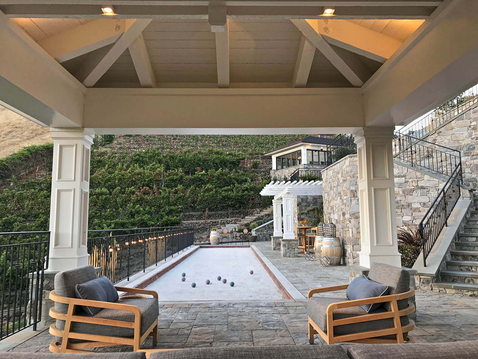 Pyramid-roofed white pergola covers lounge area at head of bocce ball court. Beyond, a slatted wood pergola, stone staircases and wrought-iron railings frame a view of the vineyard