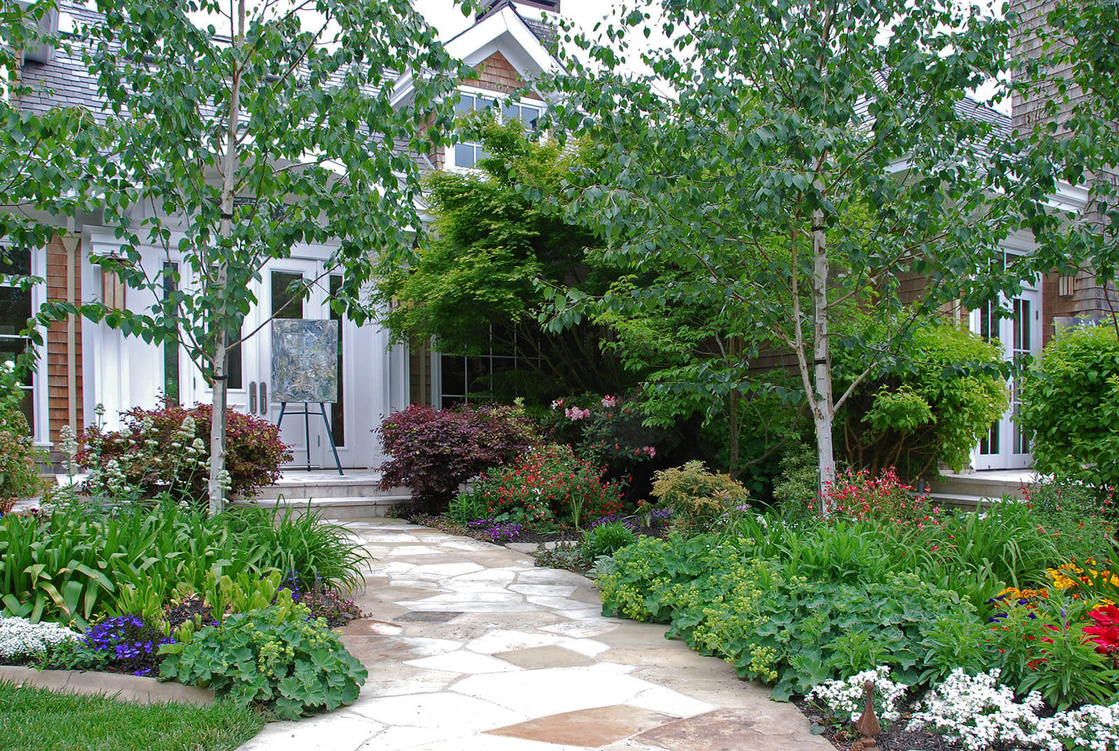 vibrant healthy garden beds with flowering plants, and dogwood trees parted by white stone walkway leading to abstract painting in front of doorway