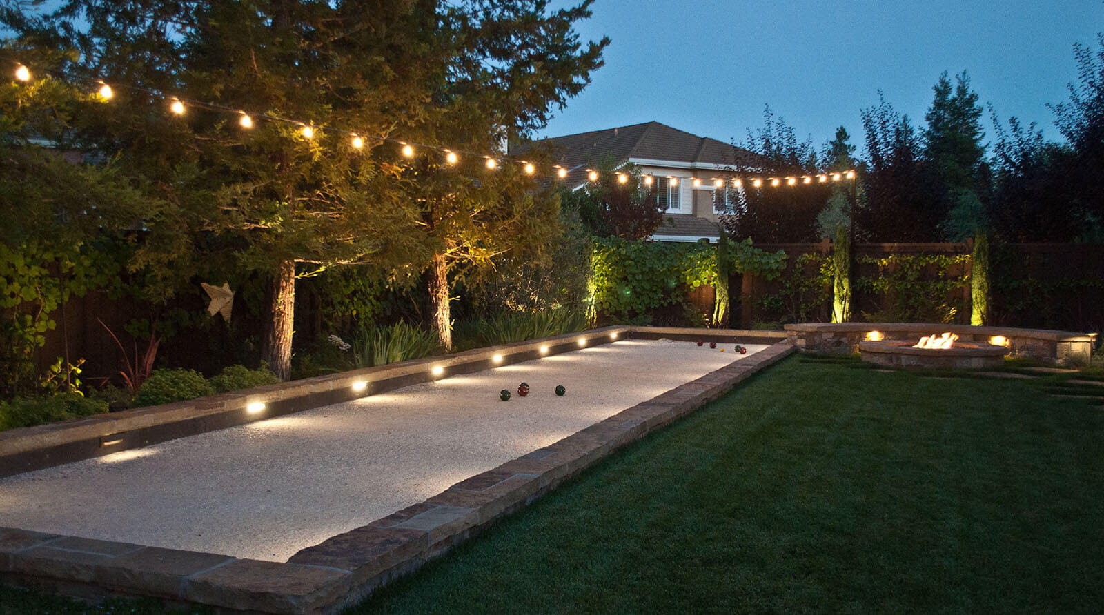 warmly lit, stone-lined bachi ball court, next to manicured lawn and fireplace with accent lighting under trees