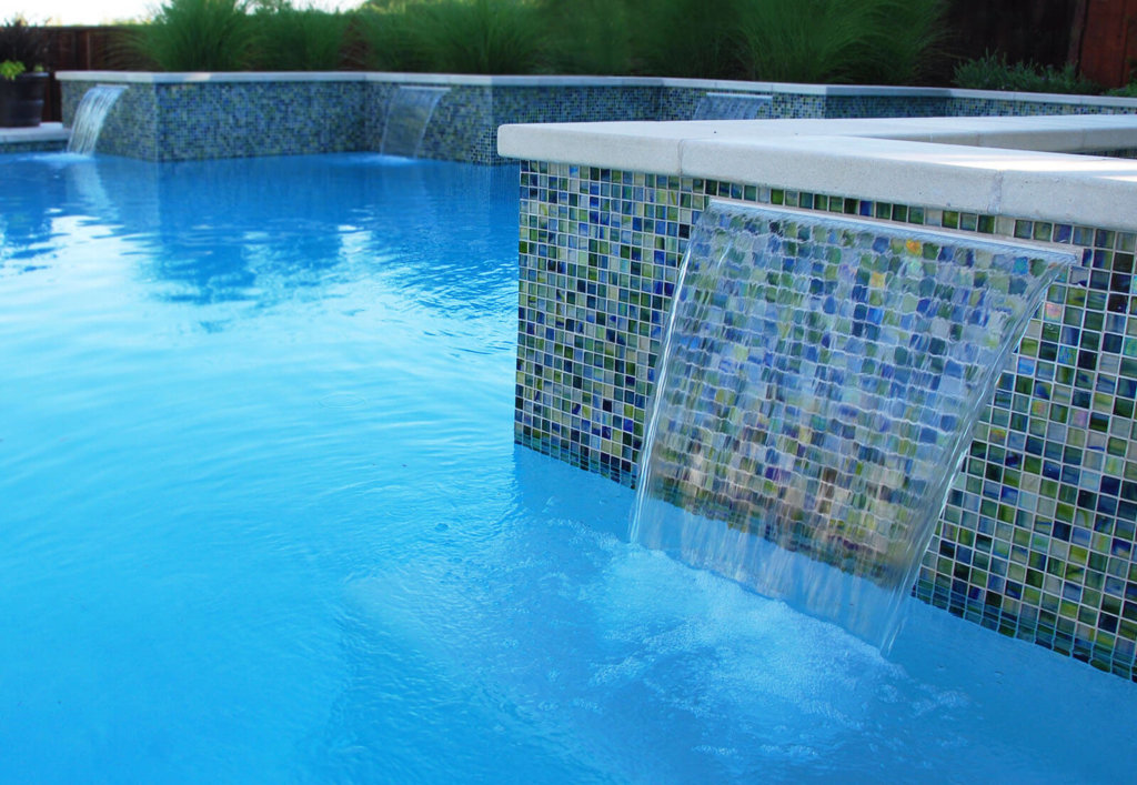 Four elevated glassy waterfalls with colorful blue-green tile mosaic backdrops topped with gray stone, flowing into a deep blue swimming pool below