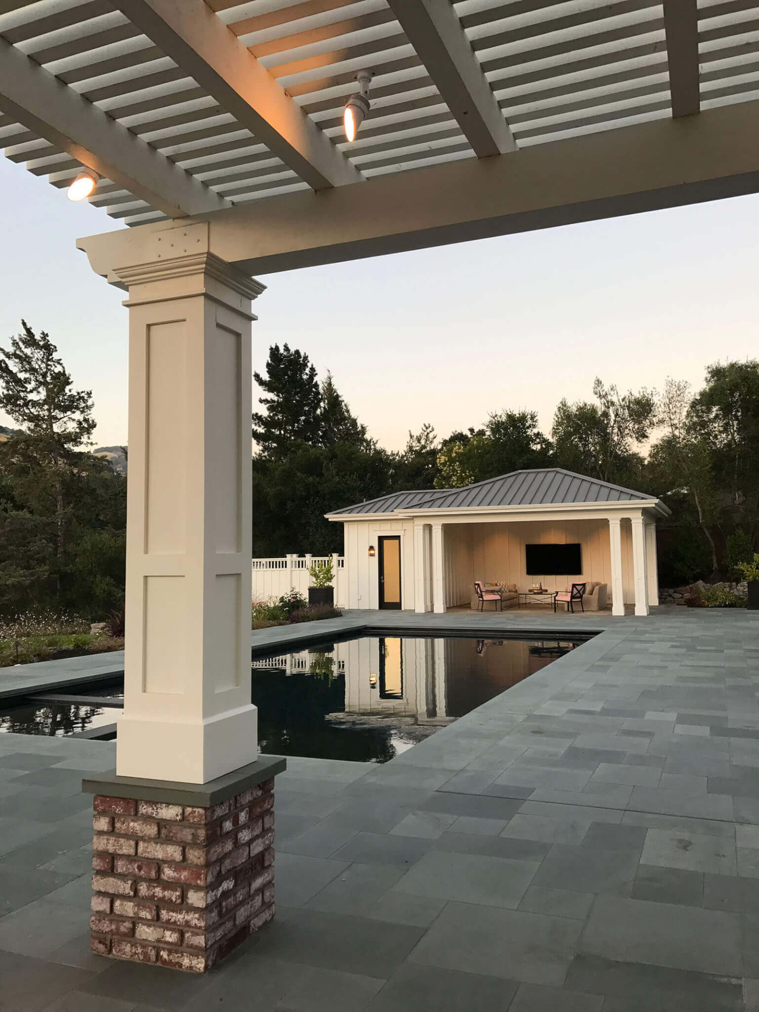 Covered lounge as seen through the posts of a matching wood pergola with brick detailing, on a bluestone patio with seamless contemporary pool and spa