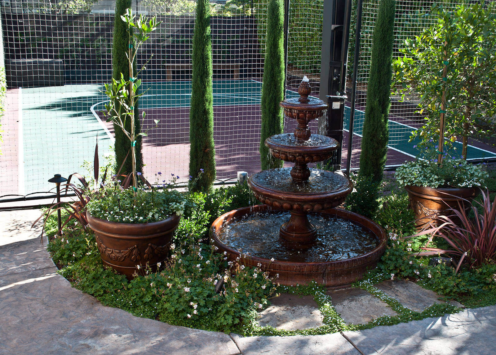 Beautiful dark brown ceramic fountain with potted trees and greenery next to private basketball court