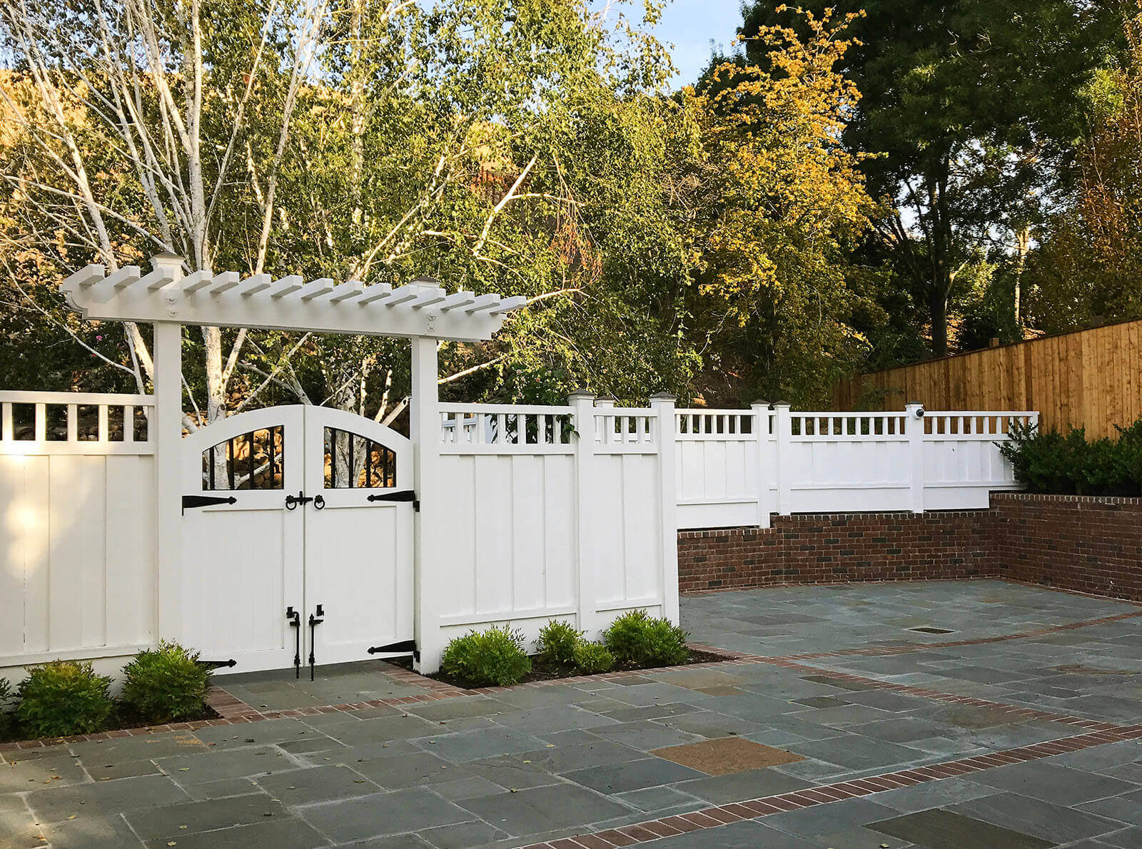 Tall white fence with double gate and arbor, wrought iron details overlooks a bluestone and brick patio