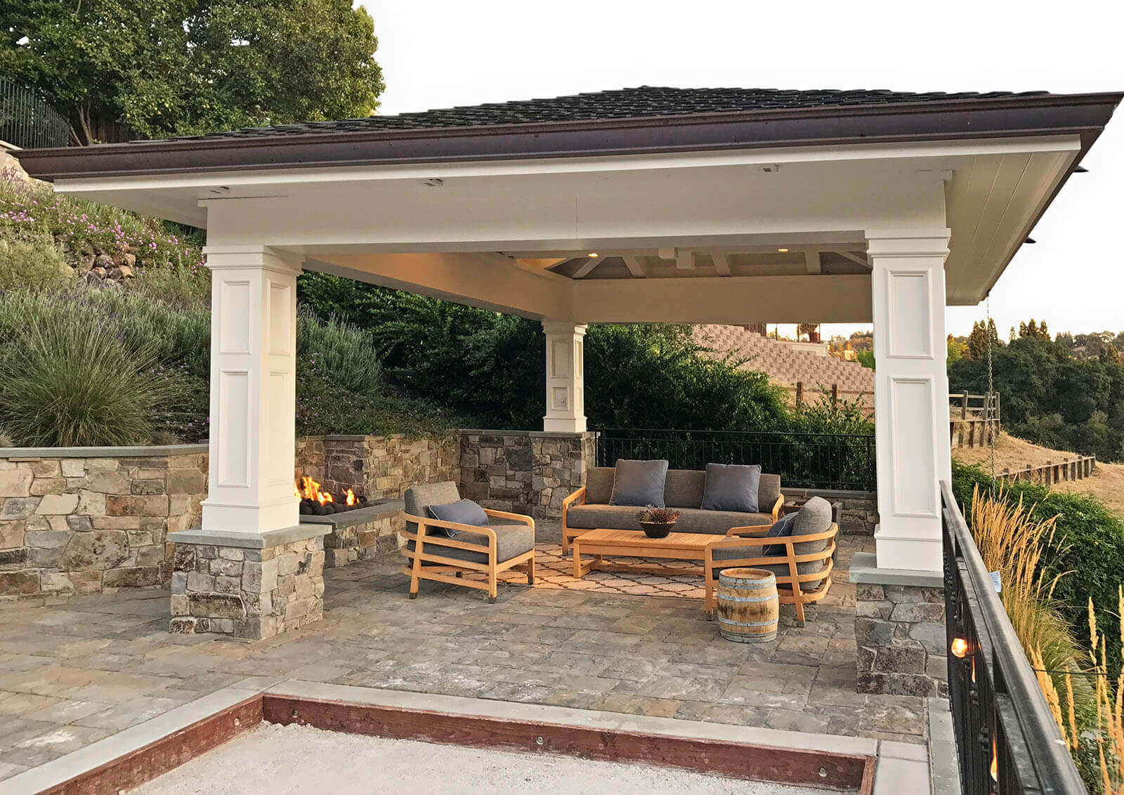 Shingled pergola shades a Sonoma-style bocce lounge on a stone terrace with hillside view