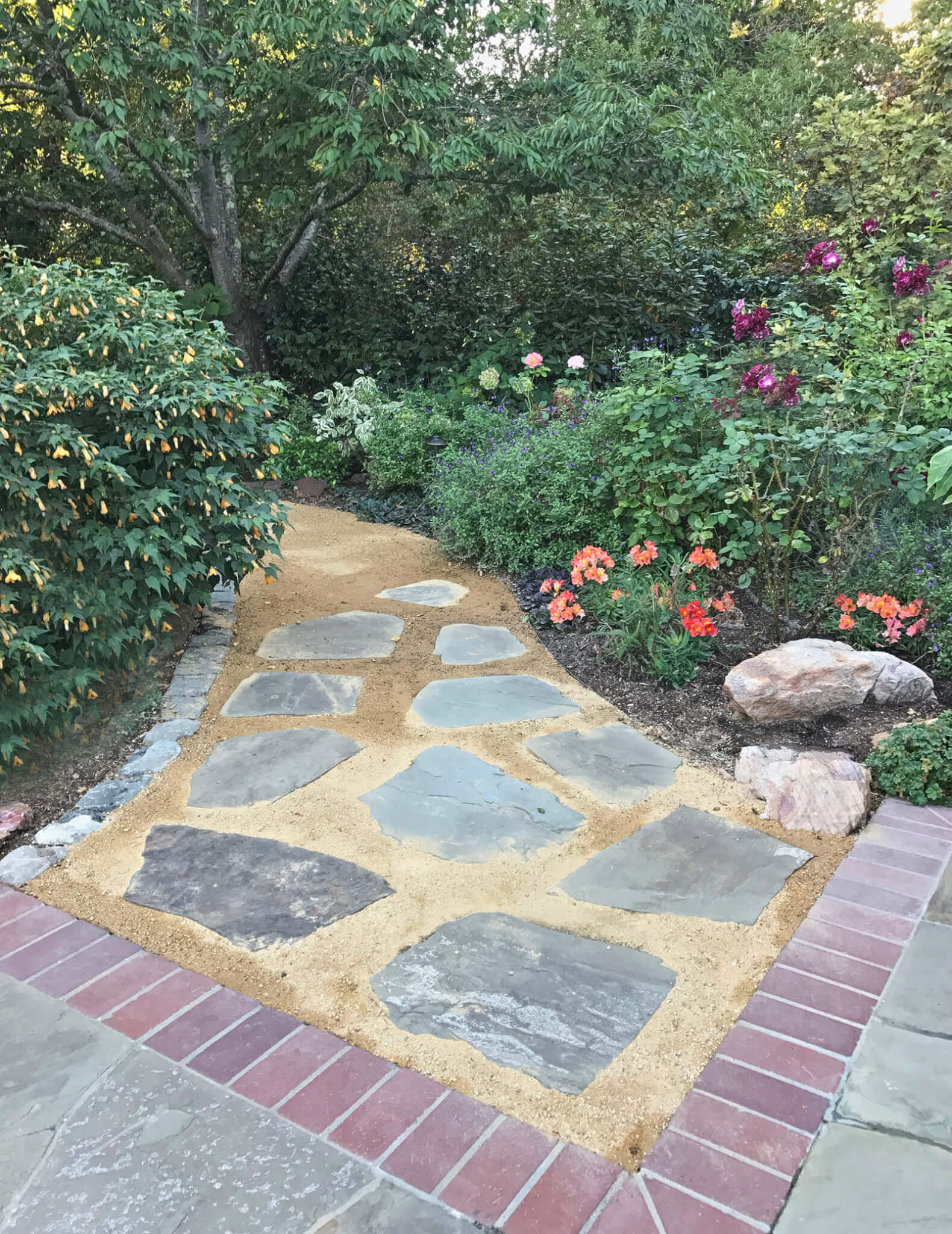 Sand and gravel walkway with laid stone leading to brick-lined stone tile patio