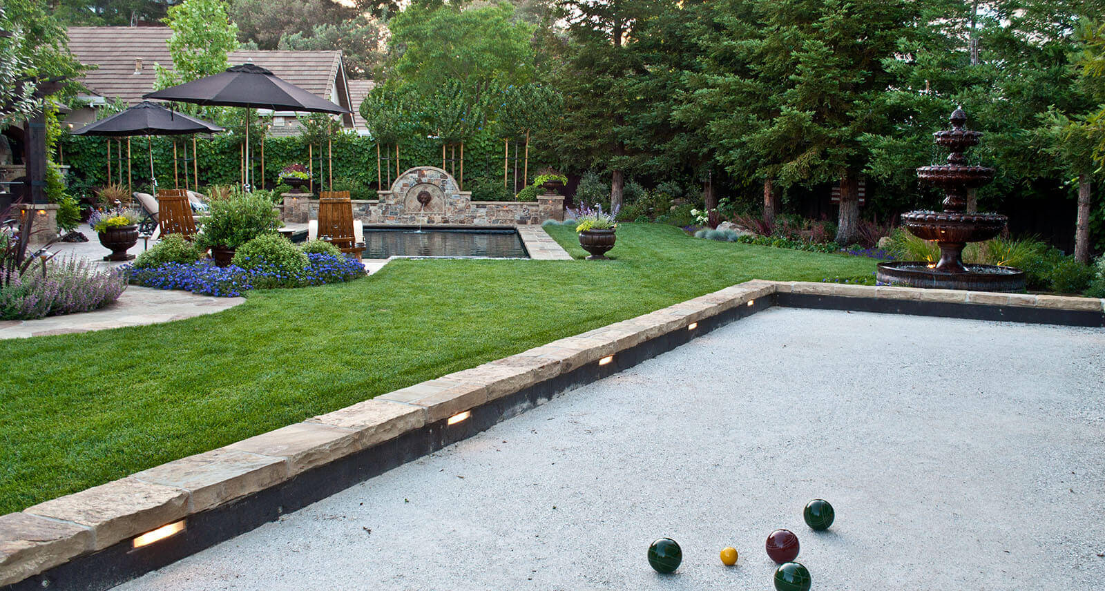 Angled view from stone-lined and lit bachi ball court, looking across lawn to pool and umbrella shaded stone patio