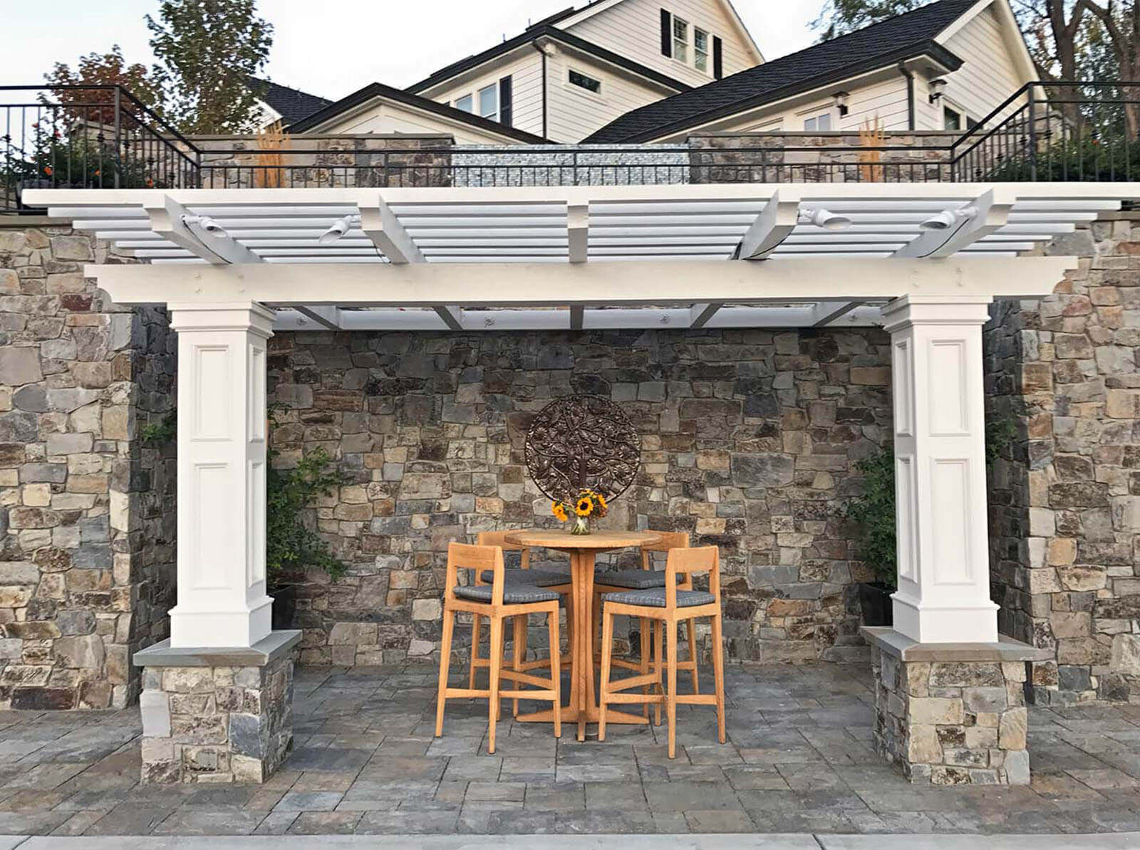 Classic white wood pergola with stone detailing set against stone wall shelters a small seating area on the lower terrace