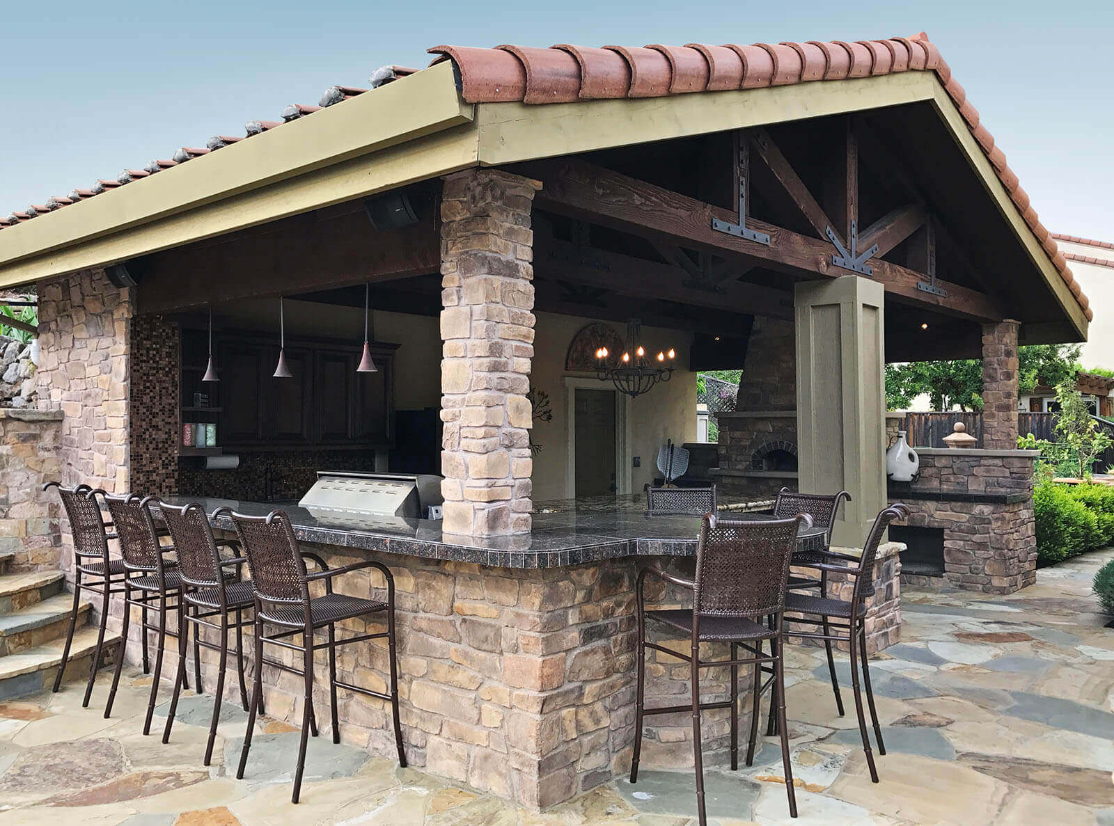 Pergola with mission-style tile roof covers outdoor bar, dining, bbq and fireplace with stone detailing on flagstone patio