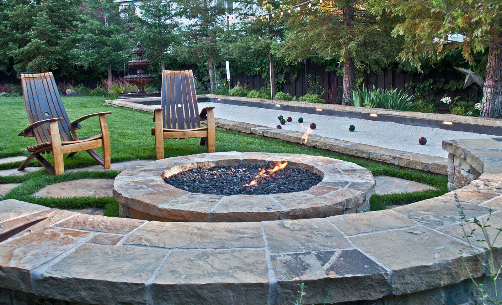Rounded stone-lined river rock fireplace with rustic wooden lawn chairs and stone wrapped seating overlooking stone-lined lit bachi ball court
