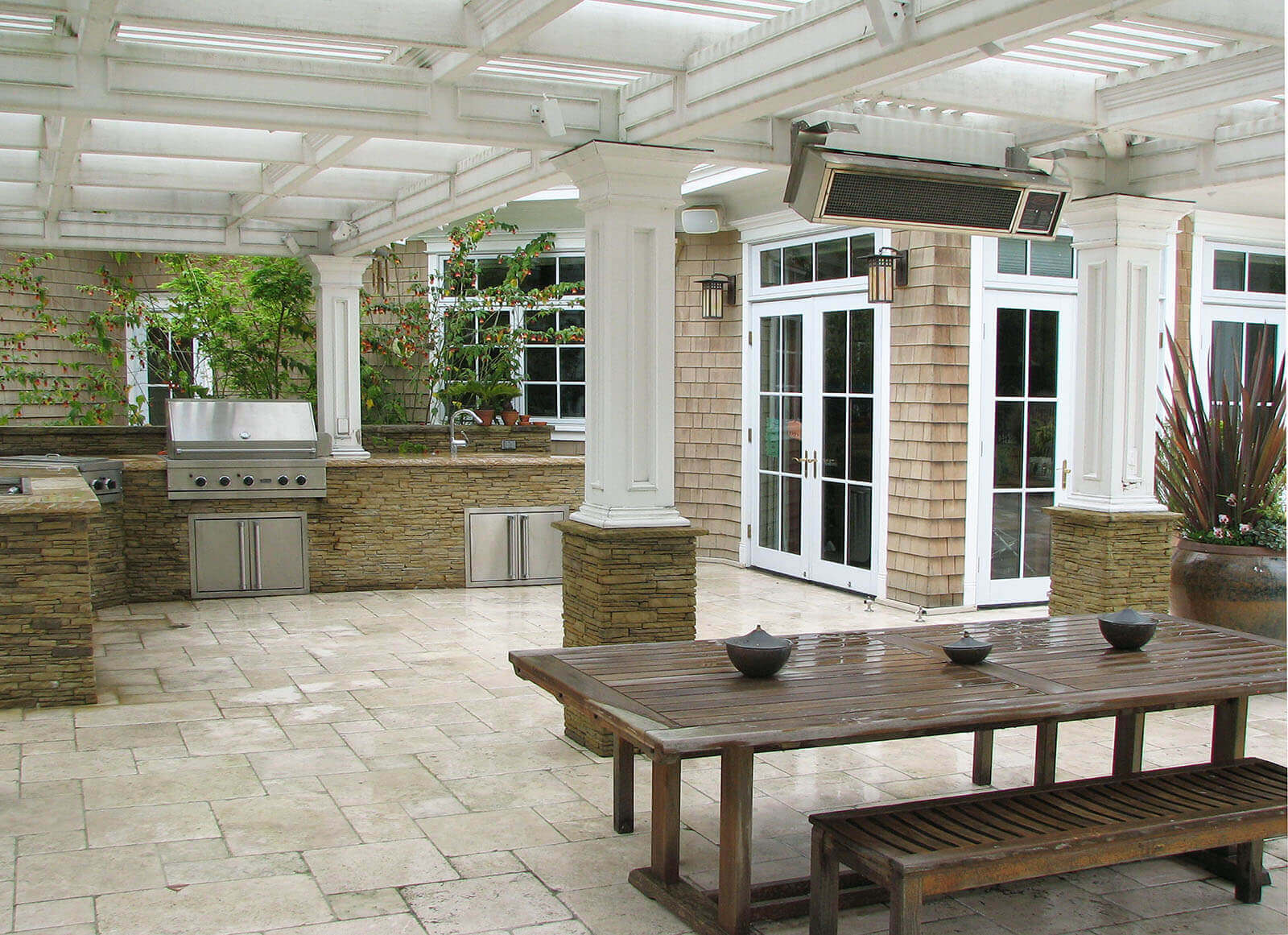 Expansive white pergola with stone column detailing shelters an outdoor kitchen and dining area with seasonal heating