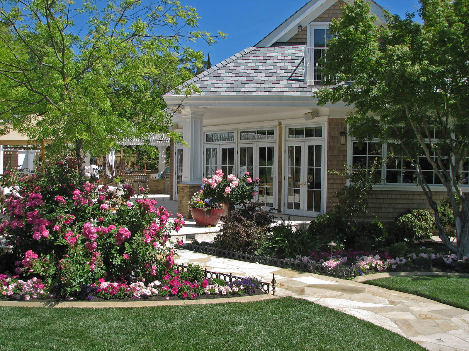 manicured lawn with stone tile path with wrought iron path borders of flowering garden patches neatly placed around a raised stone patio with extending roof with white column