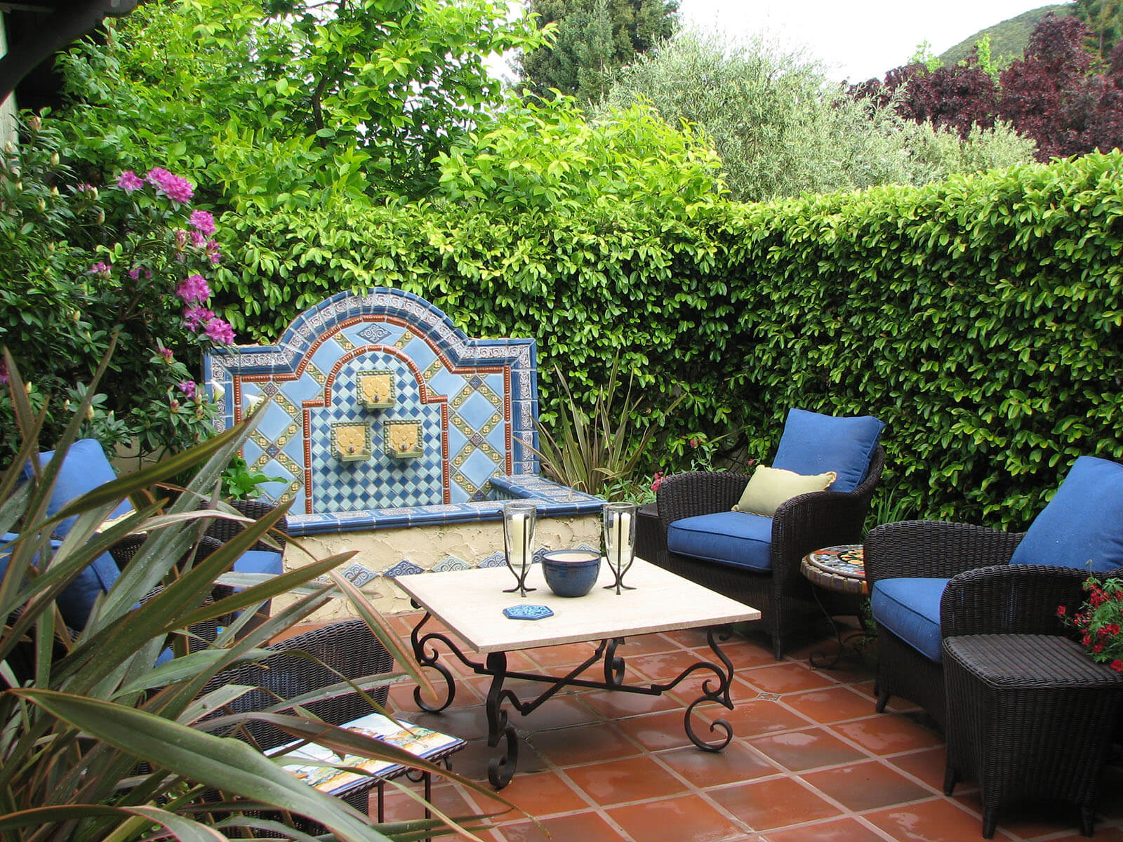 greenery walls hiding a quaint brick patio with blue and red mosaic fountain and outdoor dining set