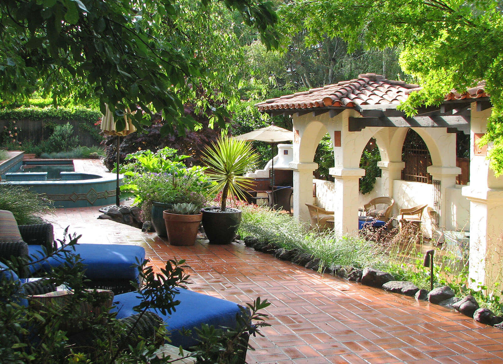 Pretty white plaster Spanish style arches and mission tile roofed seating areas with outdoor furniture in a brick tile patio and staged garden areas shrouded with trees