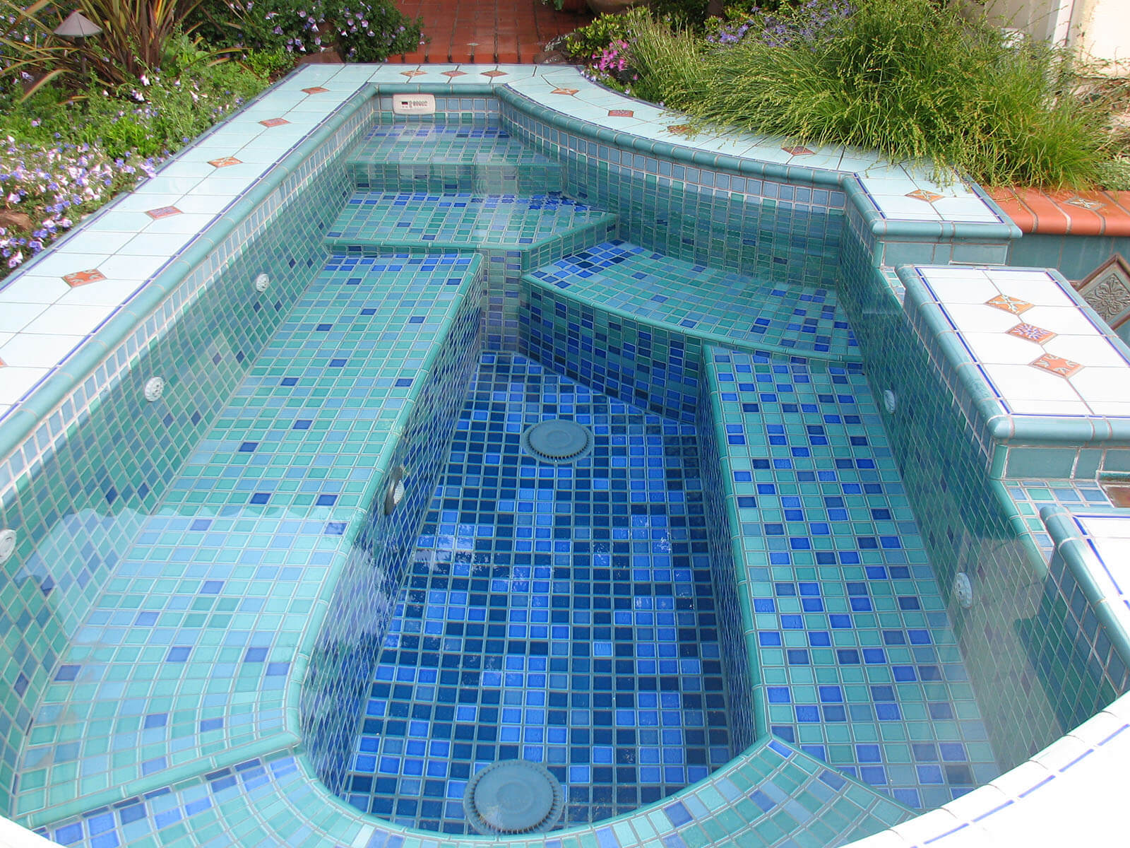 Custom shaped Jacuzzi with artistic blue mosaic tiling with white and tan topped mosaic borders flowing into pool below.