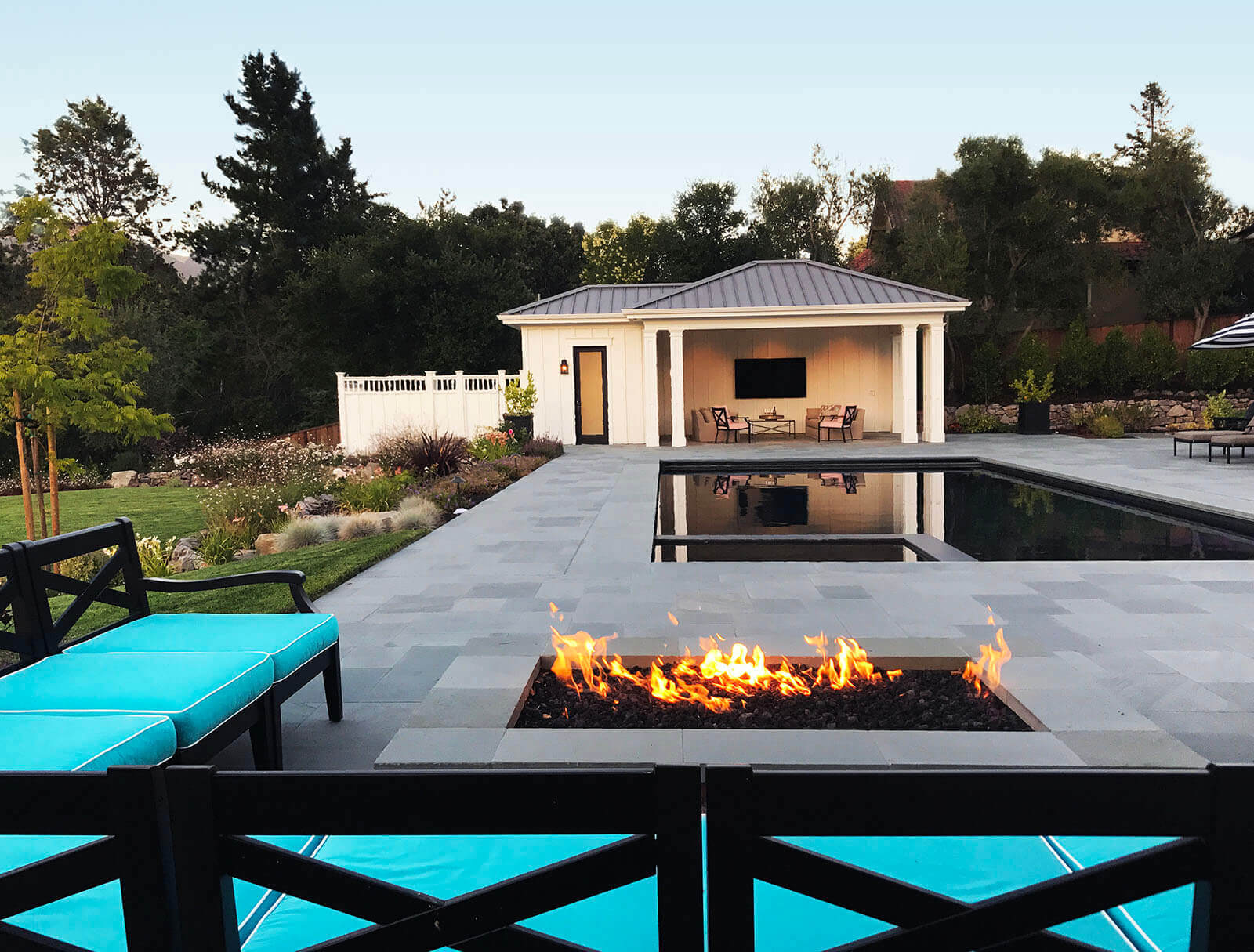 Black wooden chairs with light blue accent cushions wrapping around a short stone fireplace with black rock coals, looking on to a tranquil dark pool and jacuzzi with covered lounging area in the back