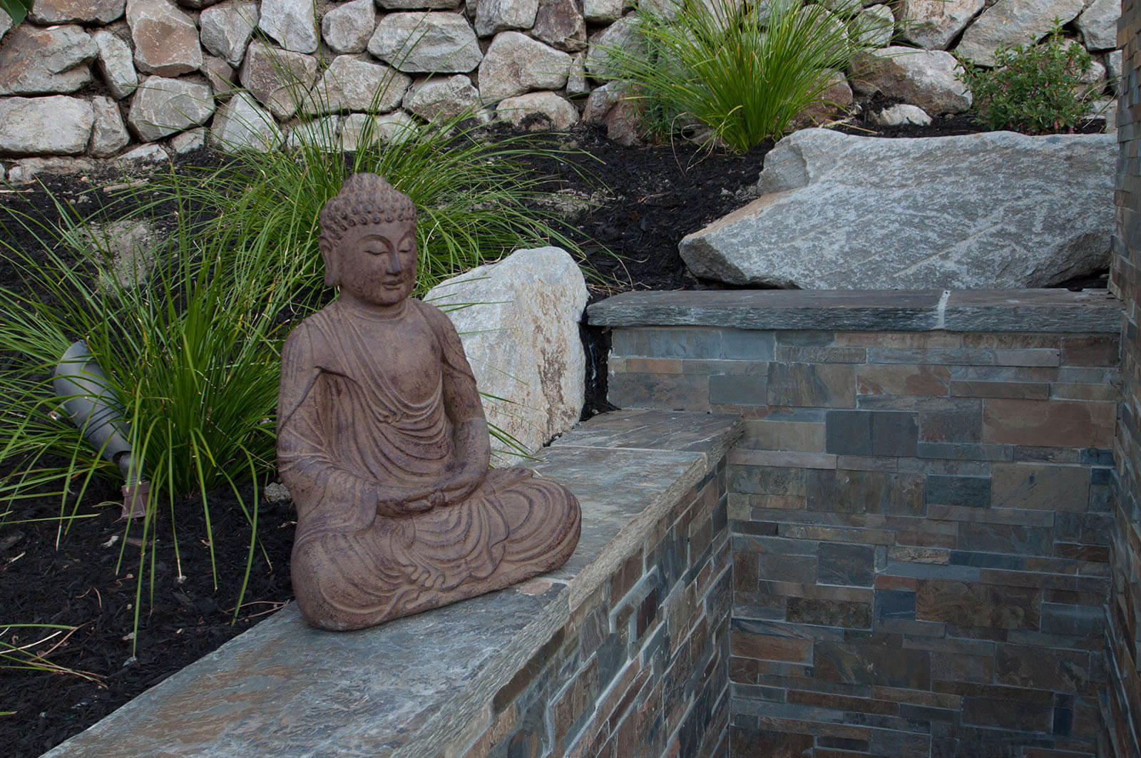 stone tiled nook in garden with meditating Buddha statue with dark soil and ornamental grasses below rock wall