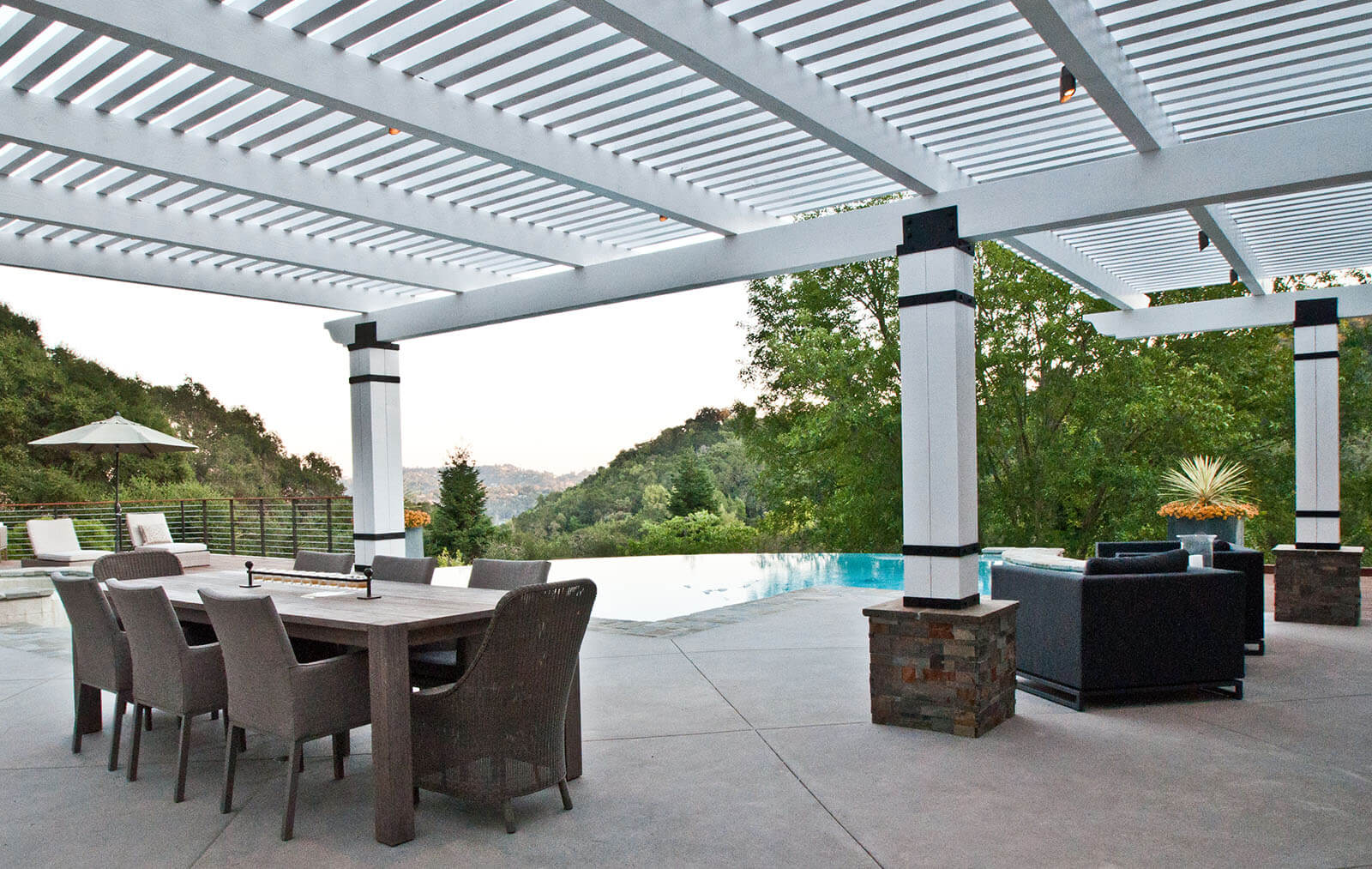 Large slat roofed, stone tiled patio, with white columns and wrought iron fixtures with stone tile bases, washed wood dining set and a view of the edgeless pool overlooking green lush valley landscape
