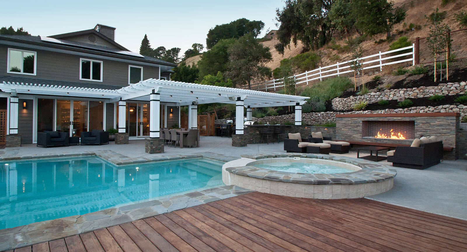 Open stone patio, half-covered by a white slatted pergola, with a nearby custom shaped aquamarine pool with elevated stone wrapped jacuzzi seating with view of broken glass fireplace, and nearby dark wood deck connecting to the pool