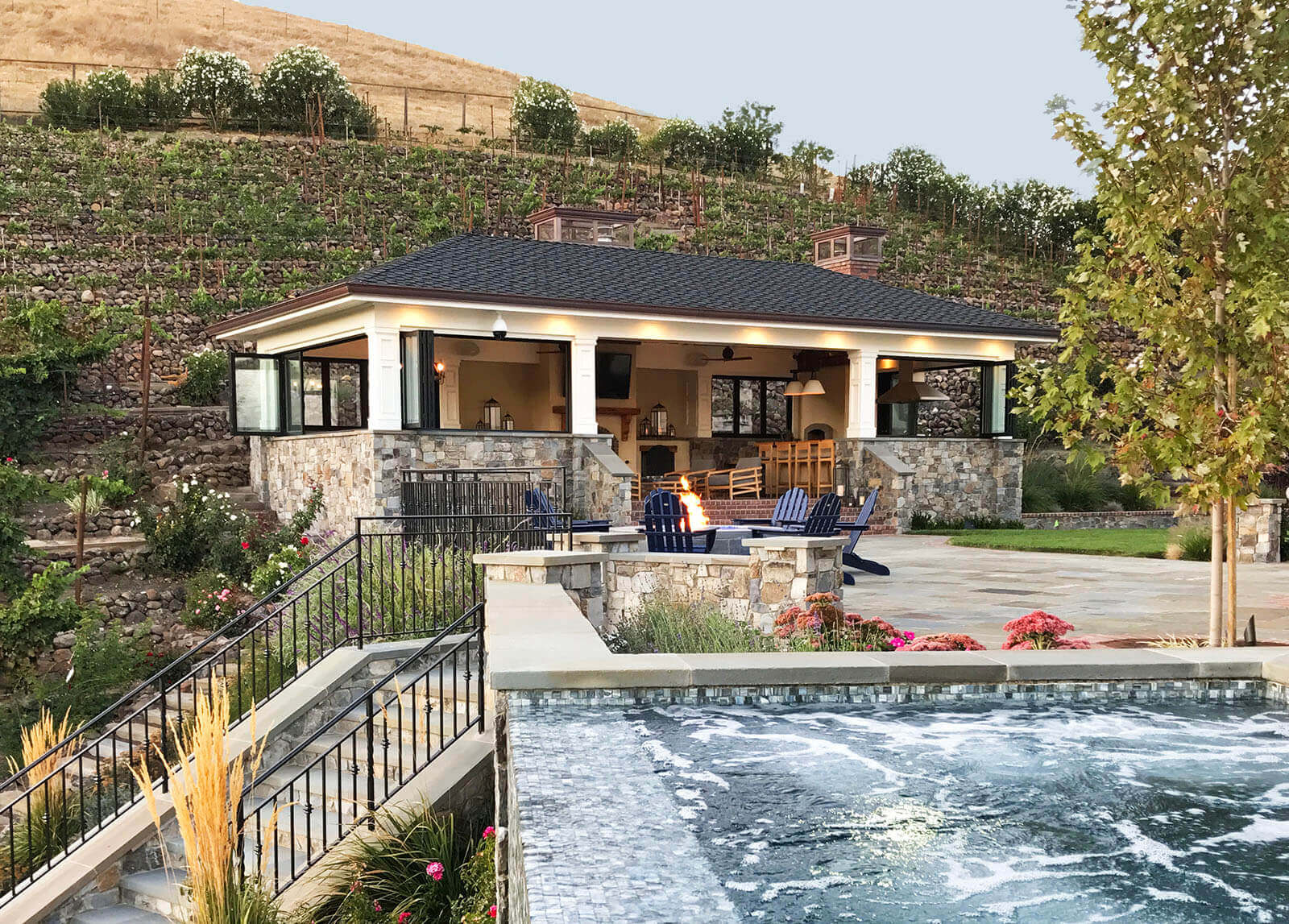 Flowing tile fountain with wrought iron railing on stone terrace, round stone fire pit with Adirondack chairs, and cabana set into hillside
