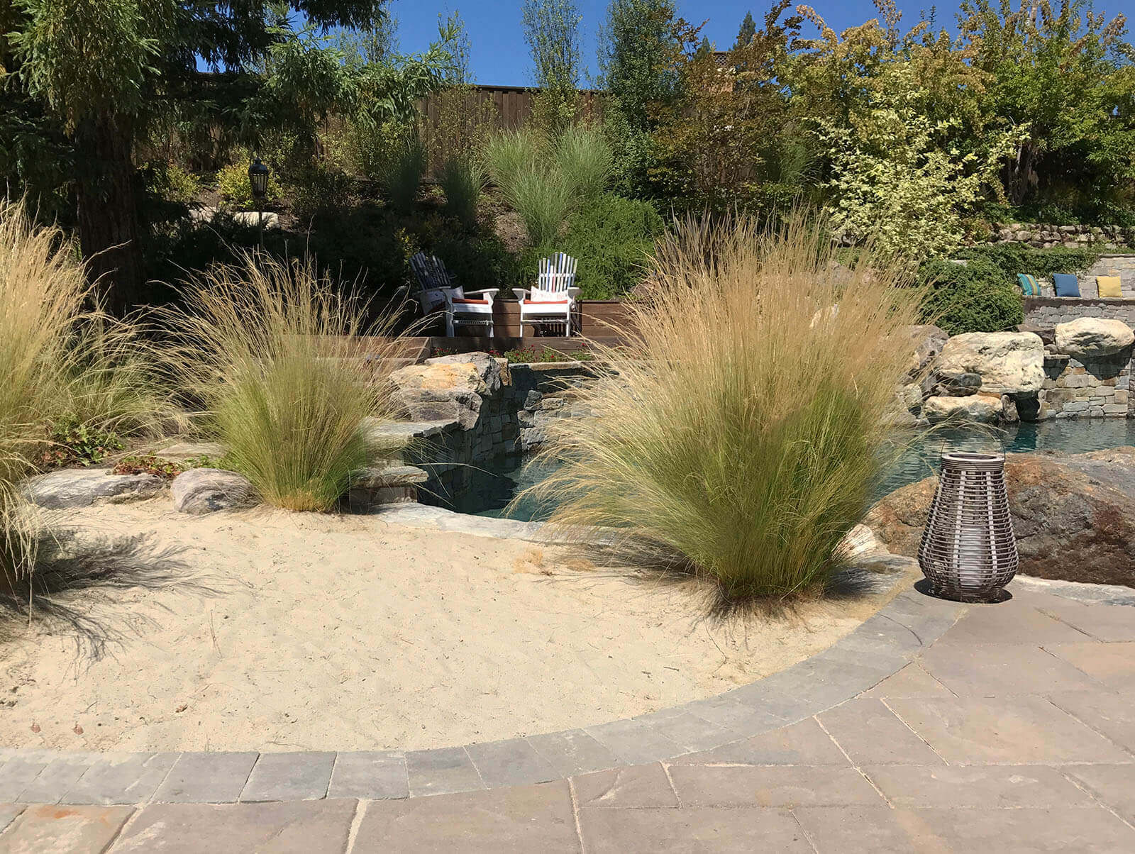 Rock-lined slate stone patio leading to ornamental grass decorated sandy beach area on edge of pool and walkway to raised wooden deck with lounge chairs