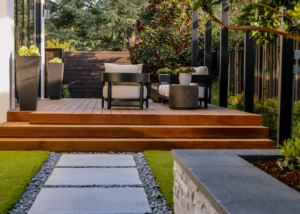 Elegant wood deck with full-length steps meets with contemporary concrete pavers, faux turf and river rock detailing
